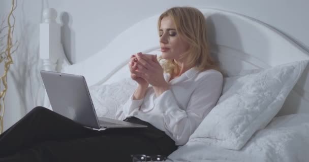 Empresária trabalhando em laptop goza de uma xícara de café quente antes de dormir depois de um longo dia difícil. Mulher de negócios relaxa na cama depois de um longo voo. Conceito de negócio. Ângulo lateral de perto. Prores 422 — Vídeo de Stock