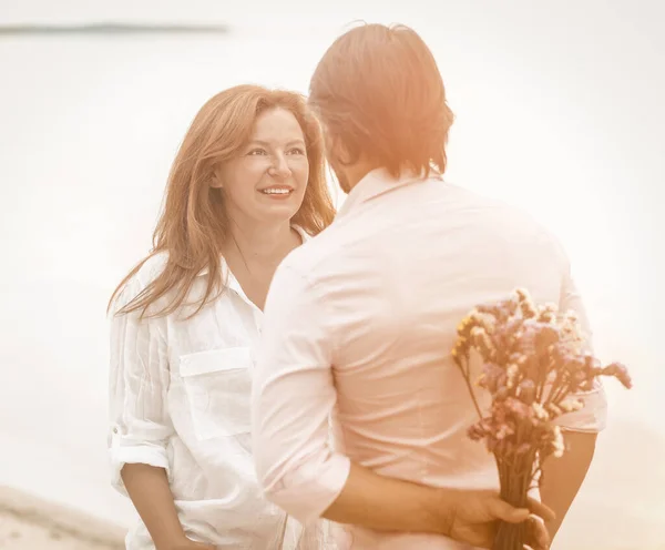 Dating of happy mature couple on seashore. Man about to give bouquet of flowers standing with his back in foreground. Selective focus on smiling mid-aged Caucasian woman looking at him. Toned image — Stock Photo, Image
