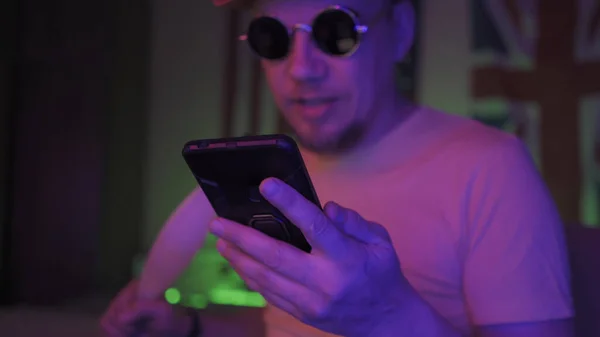 Hookah smoker using mobile phone. Handsome Caucasian Man in sunglasses sits in neon backlit. Selective focus on male hand holding phone in foreground — Stock Photo, Image