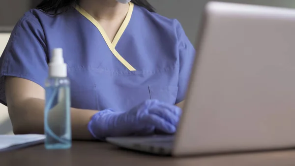 Zbliżenie lekarza piszącego na komputerze. Skup się na tułowiu medyka w niebieskim mundurze ochronnym i rękawiczkach pracujących w biurze. Widok dolny — Zdjęcie stockowe