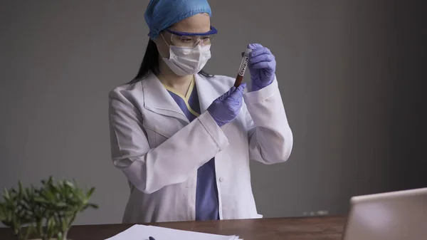 Asistentul de laborator examinează tubul de testare cu sânge pentru testul coronavirusului. Femeie medic care ține tubul de testare cu inscripția Covid 19 în mână și se uită cu atenție la el — Fotografie, imagine de stoc