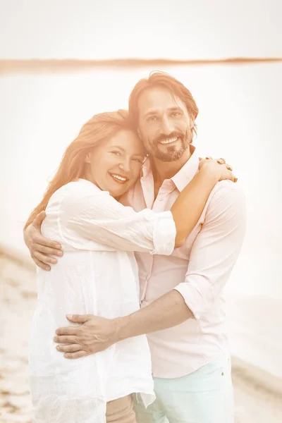 Happy couple hugs on the beach. Smiling man and woman in white clothes looking at camera while standing embracing on sea coast against backdrop of water. Toned image — Stock Photo, Image