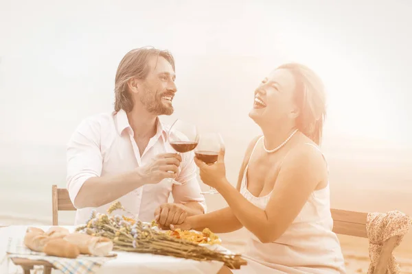 Счастливые мужчина и женщина пьют вино в пляжном кафе. Прекрасная пара смеется, держась за руки и наслаждаясь жизнью, сидя вместе на улице. Романтическая концепция среднего возраста. Тонированное изображение — стоковое фото