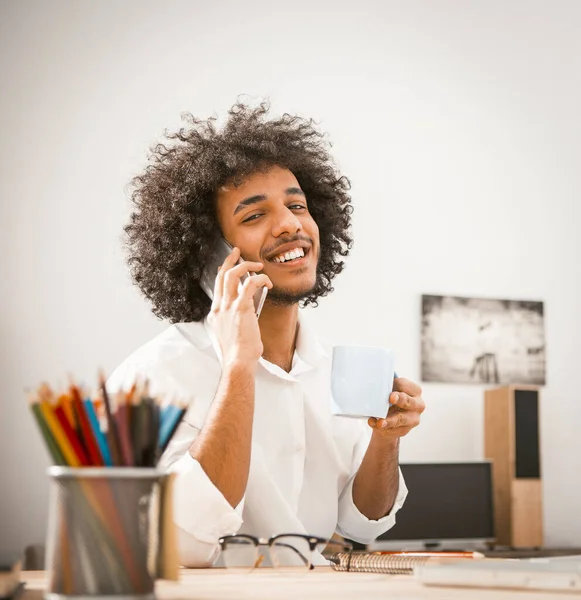 Affascinante ragazzo arabo sorrisi dentini parlando su smartphone. L'uomo positivo tiene la tazza di bevanda calda e guarda la fotocamera mentre è seduto sul posto di lavoro di casa. Concetto pausa caffè. Immagine tonica — Foto Stock