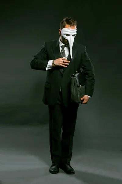 Masked businessman puts large wad of money in his breast pocket holding briefcase under his arm. Man in fancy white masks full-length stands on gray background. Corruption concept. Toned image — Stock Photo, Image