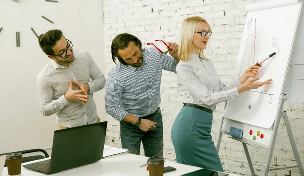 Ελκυστική επιχειρηματίας δείχνει διαγράμματα σε μια λευκή σανίδα gesturing με ενθουσιασμό, ενώ οι άνδρες συνάδελφοί της αποσπούν την προσοχή θαυμάζοντας την ομορφιά της. Σχέδιο σεμιναρίου. Τονισμένη εικόνα — Φωτογραφία Αρχείου