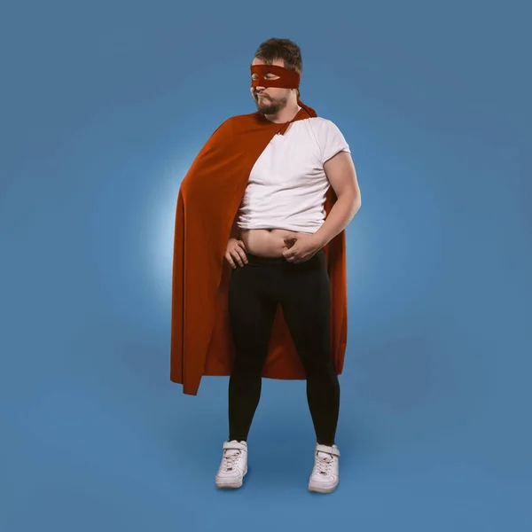 ボディポジティブなスーパーヒーローの男は冬に体重を追加しました。スーパーヒーローの衣装の悲しい男は彼のおなかの脂肪をチェックします。青い背景に隔離されています。テキストのための場所とテンプレート。太りすぎの概念 — ストック写真