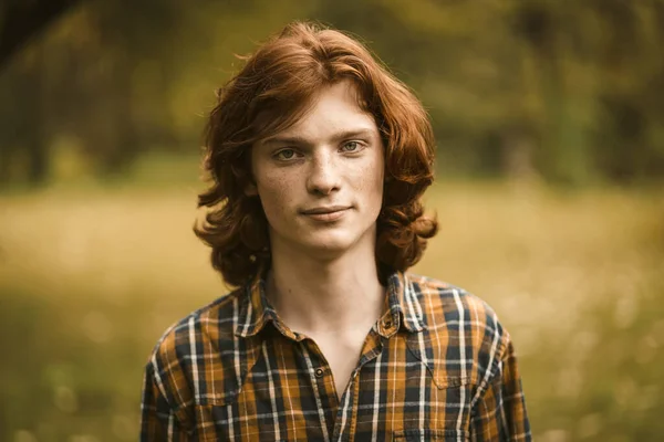 Roodharige man met sproeten poserend in de natuur. Knappe jongeman in een geruit shirt kijkt naar de camera die licht glimlacht terwijl hij tegen een groen gazon staat. Mannelijk schoonheidsconcept. Getinte afbeelding — Stockfoto