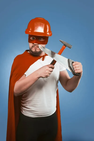 Superhero builder or repairman holds construction tools. Man in red helmet and Superhero costume crossed saw and hammer while looking at camera while standing on a denim blue background — Stock Photo, Image