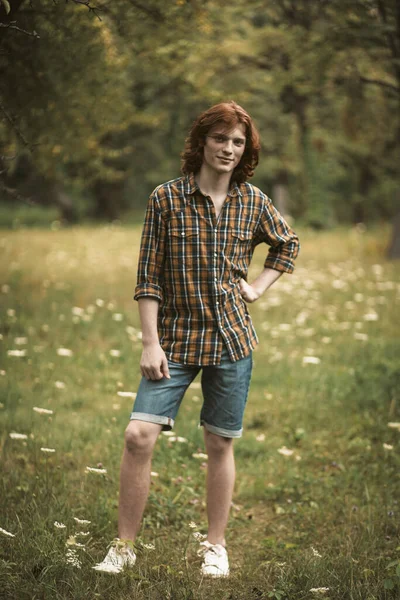 Smiling young man with long red hair posing full growth against backdrop of blooming meadow. Slim Caucasian guy wearing in checkered shirt, denim shorts and white sneakers standing looking at camera — Stock Photo, Image