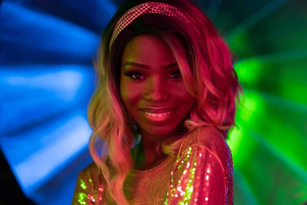 Charmante Afroamerikanerin lächelt geheimnisvoll im Neonlicht. Sexy Frau mit ombre Frisur in leuchtendem Kleid in grün-rosa und blauem Neon-Schein. Disco-Konzept — Stockfoto