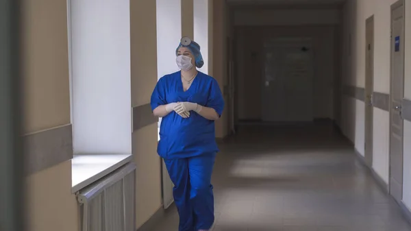 Dokter wanita atau perawat berseragam biru berjalan di sepanjang koridor rumah sakit sambil melihat jendela. Rumah Sakit Kota. Mei 2020, Brovary, Ukraina — Stok Foto