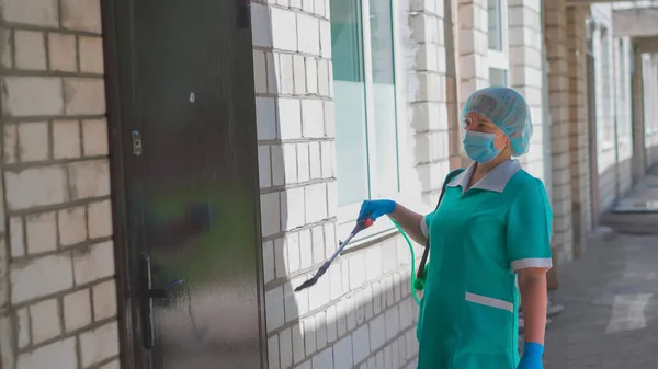 Femeia în uniformă verde dezinfectează ușa și peretele exterior al spitalului. Tratamentul medical preventiv al clădirii cu antiseptic. Spitalul Municipal. Mai, 2020, Brovary, Ucraina — Fotografie, imagine de stoc