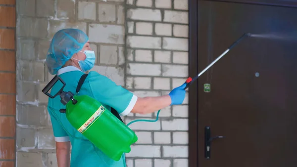 Masked woman cleaning door with liquid disinfectant using large bottle and spray. Antiseptic treatment concept. Preventive medical treatment. City Hospital. May, 2020, Brovary, Ukraine — Stock Photo, Image