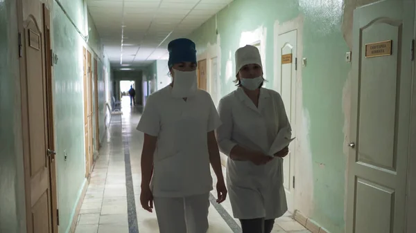 Două asistente de sex feminin mascate în haine albe care se plimbă prin sala de spital uitându-se la cameră. Interiorul spitalului în proces de reparații. Imagine abstractă defocalizată. Spitalul Municipal. Mai, 2020, Brovary, Ucraina — Fotografie, imagine de stoc