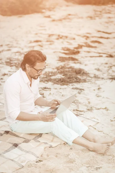 Homem criativo trabalhando com laptop sentado em uma praia de areia. Homem de meia-idade caucasiano em roupas brancas está digitando no teclado do computador, trabalhando remotamente. Imagem tonificada. Conceito freelance. Imagem tonificada — Fotografia de Stock