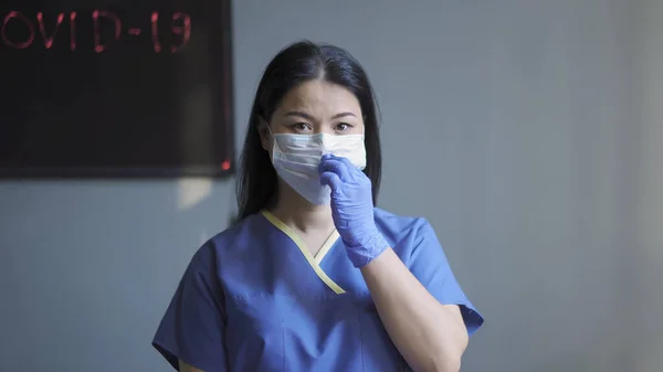 Doctorul mascat lucrează în biroul ei. Femeie asiatică în mască și uniformă de protecție privind camera împotriva peretelui gri cu tablă de neon și inscripție covid-19 pe ea — Fotografie, imagine de stoc