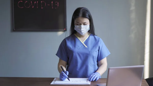 Pekerja medis menulis dengan penanda pada kertas putih. Wanita bertopeng dan seragam biru bekerja melawan dinding abu-abu dengan papan tulis neon dan tulisan covid-19 di atasnya — Stok Foto