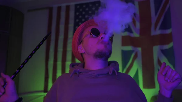 Κομψό hipster καπνίζει ένα hookah στο εσωτερικό του σπιτιού με φόντο την αμερικανική και τη Μεγάλη Βρετανία σημαίες κρέμονται στον τοίχο — Φωτογραφία Αρχείου