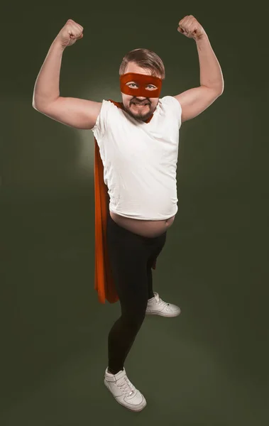 Сильный супергерой, выражающий силу. Мужчина в белой футболке, красной маске и плаще показывает бицепсы, глядя в камеру. Изолированный на зеленом фоне — стоковое фото