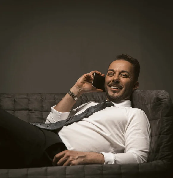 Businessman talks over mobile phone lying on sofa. Smiling man at couch talking smartphone on grey back. Close up shot. Toned image — Stock Photo, Image