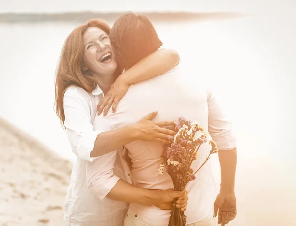 Glückliches Paar umarmt sich am Meeresufer. Lachende Frau umarmt einen Mann, der an einem Sommerabend einen Blumenstrauß hinter sich hält. Überraschungskonzept. Getöntes Bild — Stockfoto