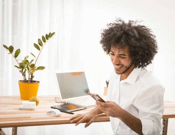 Felice giovane uomo in chat su smart phone denti sorridenti. Studente arabo o imprenditore con lunghi capelli ricci utilizzando il telefono cellulare che lavora al tavolo di legno con laptop su di esso. Immagine tonica — Foto Stock