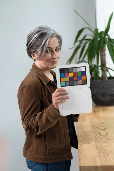 Jolie femme tient le contrôleur de couleur ou la cible spéciale pour l'ajustement de la couleur tout en se tenant dans l'intérieur du bureau. Femme aux cheveux gris montrant la palette de couleurs pour ajuster la couleur — Photo