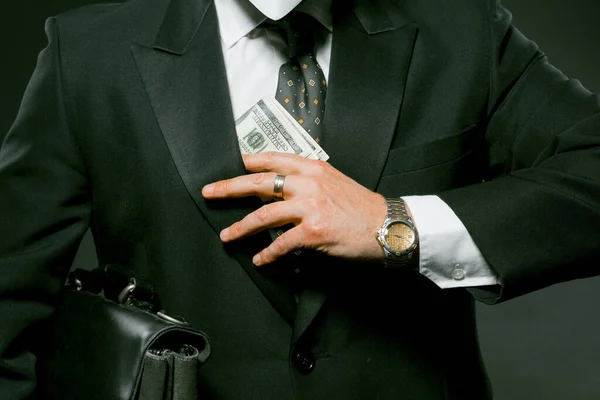 Businessman puts wad of banknote in his pocket holding black briefcase under his arm. Caucasian man wearing black suit holding money on gray background. Close up shot. Toned image — Stock Photo, Image