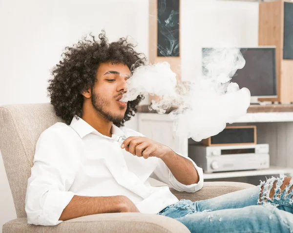 Молодой человек курит электронную сигарету дома. Араб обожает облака дыма, курящие электронные сигареты. Никотиновая зависимость. Альтернативная концепция курения. Тонированное изображение — стоковое фото