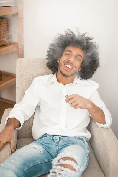 Χαμογελώντας καπνιστής άνθρωπος χαλαρώνει το κάπνισμα ένα ηλεκτρονικό τσιγάρο, ενώ κάθεται στο σπίτι πολυθρόνα. Νεαρός Άραβας εκπνέει σύννεφο καπνού κοιτάζοντας την κάμερα. Τονισμένη εικόνα — Φωτογραφία Αρχείου