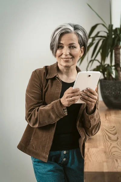 Charming woman uses digital tablet toothy smiling at camera. Mature woman wearing corduroy jacket and jeans holds tablet computer standing near wooden table or sill. Tinted image — Stock Photo, Image