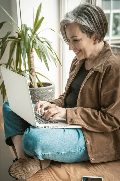 Šťastná dospělá žena si povídá na laptopu opřená o okno. Hezká dáma se široce usmívá pomocí počítače. Boční pohled. Tónovaný obrázek — Stock fotografie
