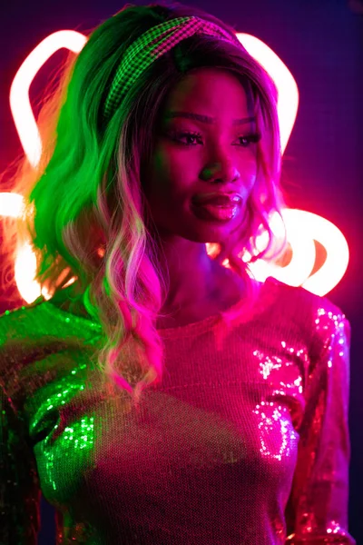 Stylish sexy African woman looking at side in pink and green lights with neon unusual backlit. Young woman with ombre hairstyle and evening make-up with long false eyelashes. Beauty concept