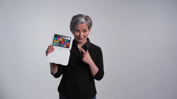 Mujer adulta de pelo gris mostrando placa de color checker y baile feliz con expresiones faciales posando para fotógrafo en estudio sobre fondo blanco en ropa casual camisa negra y jeans. Prores 422 — Vídeos de Stock