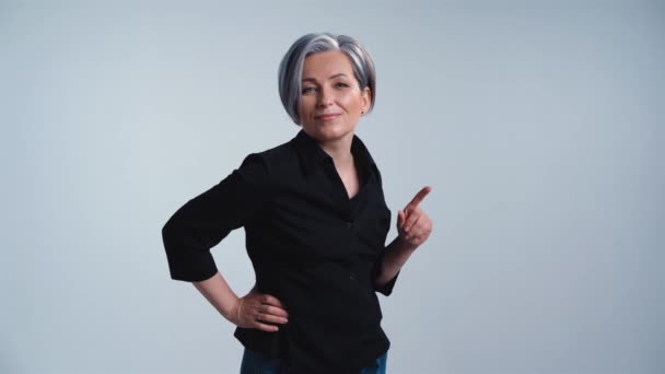 Gammal grå hår kvinna visar ansiktsuttryck med ett finger upp poserar på kameran i studio på en vit bakgrund i casual kläder svart skjorta och jeans. Prores 422 — Stockvideo