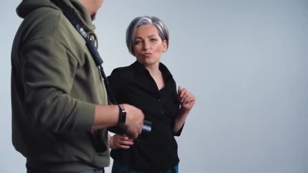 Fotograf erklärt reifen Modell erwachsene Frau mit grauen Haaren, wie man für Fotografen im Studio auf weißem Hintergrund posiert in legerer Kleidung schwarzes Hemd und Jeans. Prores 422 — Stockvideo