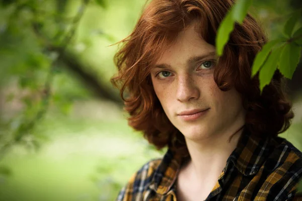 Red-haired man posing in sunny day outdoors. Young foxy guy in a plaid shirt looks at camera standing against on green floral background. Male beauty concept — Stock Photo, Image