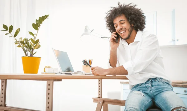 Sidovy över ung entreprenör eller student pratar mobiltelefon när du arbetar hemifrån. Charmig arabisk kille chattar på smarttelefon med vänner eller kunder. Tonad bild — Stockfoto