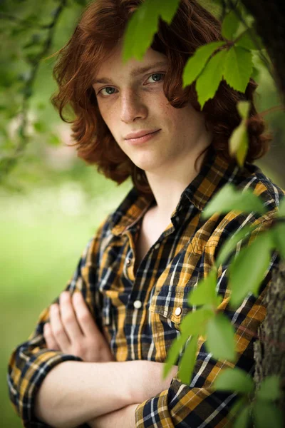 Foxy freckled guy looks at camera standing crossed hands. Red-haired young man with long hair wearing in a plaid shirt stands among green foliage. Mysterious masculine beauty concept