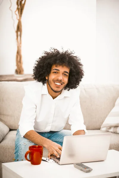अरब छात्र उस पर लाल कप के साथ कॉफी टेबल पर लैपटॉप काम करता है। सुंदर आदमी घर सोफे पर बैठे मुस्कुराता है। गृह शिक्षा अवधारणा। रंगीन छवि — स्टॉक फ़ोटो, इमेज