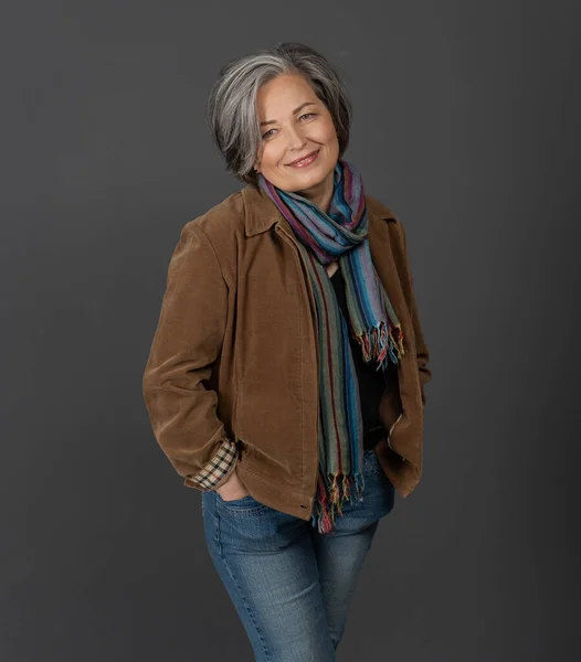 Ganska gråhårig kvinna i leende poserar på grå bakgrund. Elegant kvinna i jeans som står med händerna i fickorna — Stockfoto