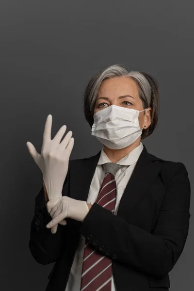 Femeie de afaceri în mască medicală care pune mănuși albe de unică folosință. Femeia matura inteleapta respecta masurile de siguranta datorate epidemiei de virus — Fotografie, imagine de stoc