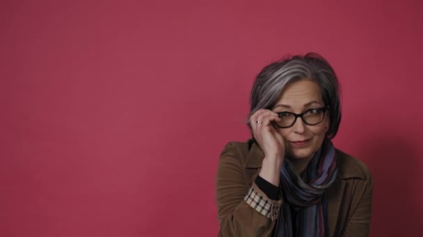 Adorable modelo maduro de pelo gris en jeans posando sobre fondo rosa. Mujer bastante madura corrige las gafas de los ojos y mirar en su teléfono inteligente. Prores 422 — Vídeos de Stock