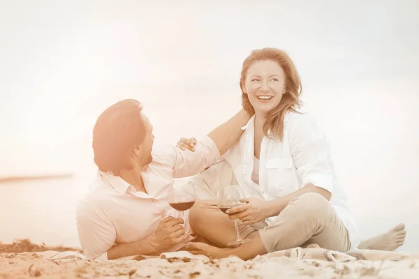 Pareja feliz disfrutar de charlar entre sí, pasar tiempo juntos. Hombre y mujer maduros beben vino abrazándose y riendo mientras están sentados en la orilla arenosa cerca del agua. Vacaciones por mar. Imagen teñida — Foto de Stock