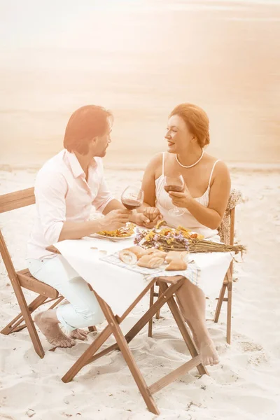 Vackra par dricker vin som håller hand medan de sitter vid bordet på strandcaféet barfota. Gammal man och kvinna vilar på sandkusten mot på havet eller havet bakgrund. Tonad bild — Stockfoto