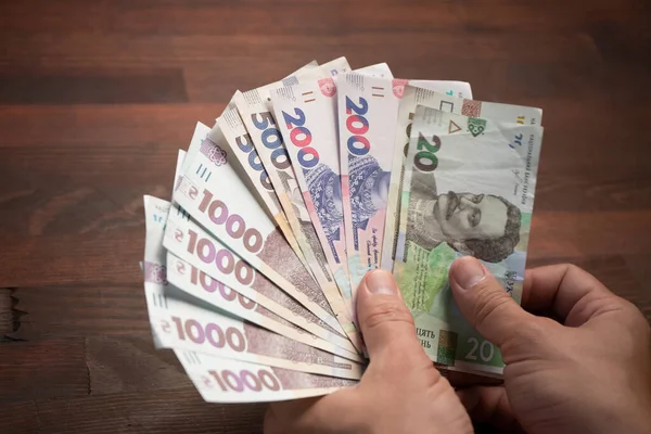 Male hands holding banknote fan. Caucasian man has pack of new Ukrainian hryvnias against of wooden table background. Close up shot. Cash money concept