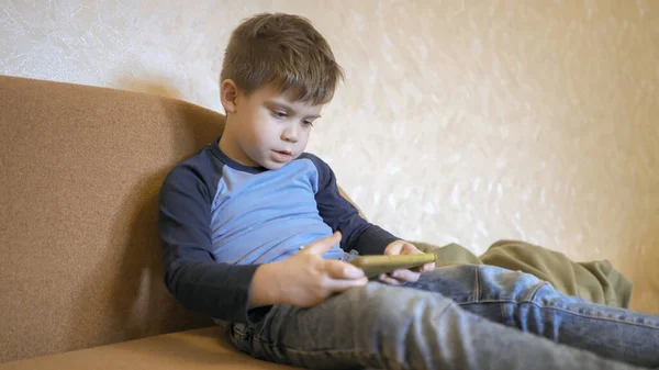 Aburrido niño utiliza el teléfono celular mientras está sentado solo en casa en el sofá. Niño preescolar caucásico mirando la pantalla del teléfono. Concepto de adicción a la dopamina — Foto de Stock