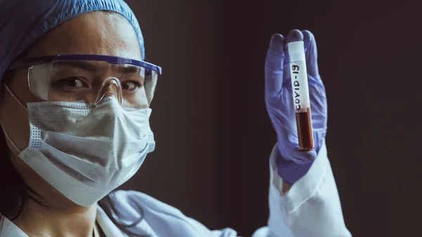 A funcionária do laboratório tem um tubo de sangue a olhar para a câmara. Mulher asiática em máscara protetora e óculos de proteção em fundo cinza. Conceito de vacina. Um tiro de perto. Imagem matizada — Fotografia de Stock