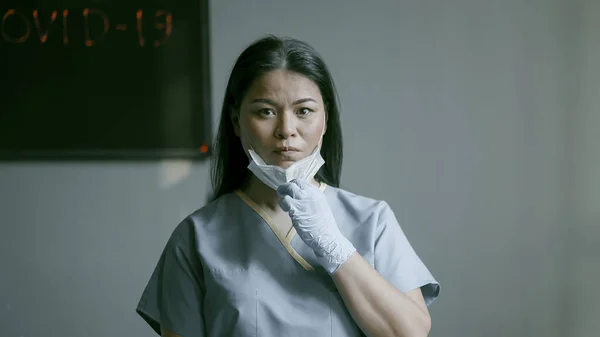 Frumoasa femeie asiatică și-a deschis fața îndepărtându-și masca de față și uitându-se serios la cameră. Femeie medic stand în birou pe fundal de perete gri. Imagine tonată — Fotografie, imagine de stoc